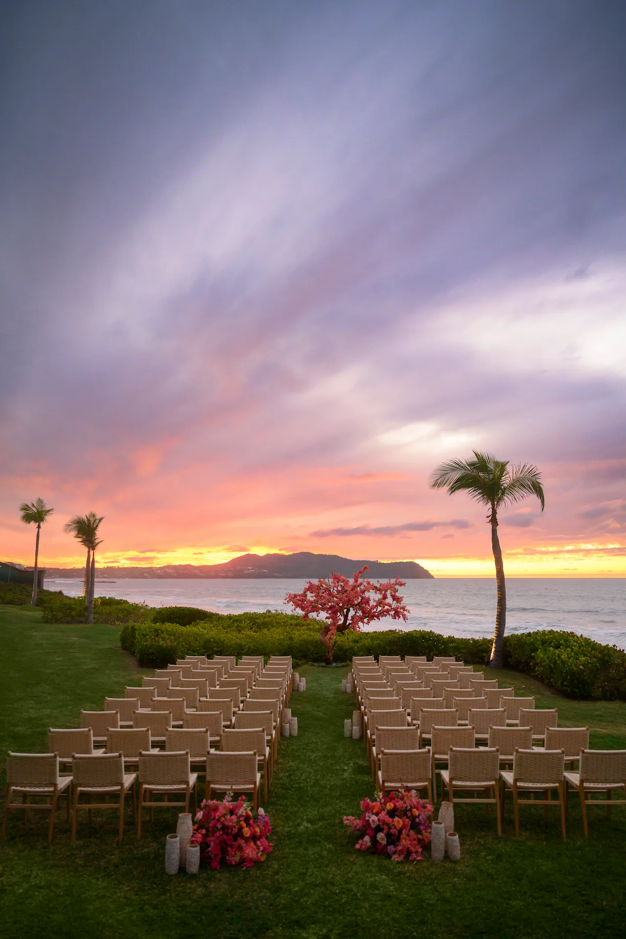 Configuración de boda al atardecer con altar de flores rosas, pasillo rodeado de faroles y vista al mar enmarcada por palmeras.