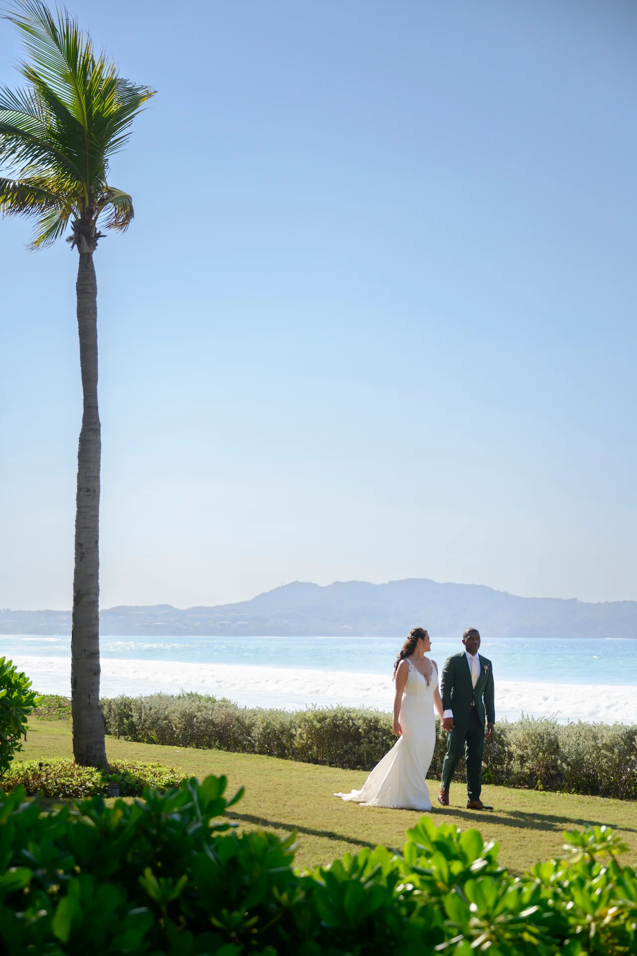 Novia y novio tomados de la mano cerca del océano con palmeras, vista a la montaña y cielo azul claro.