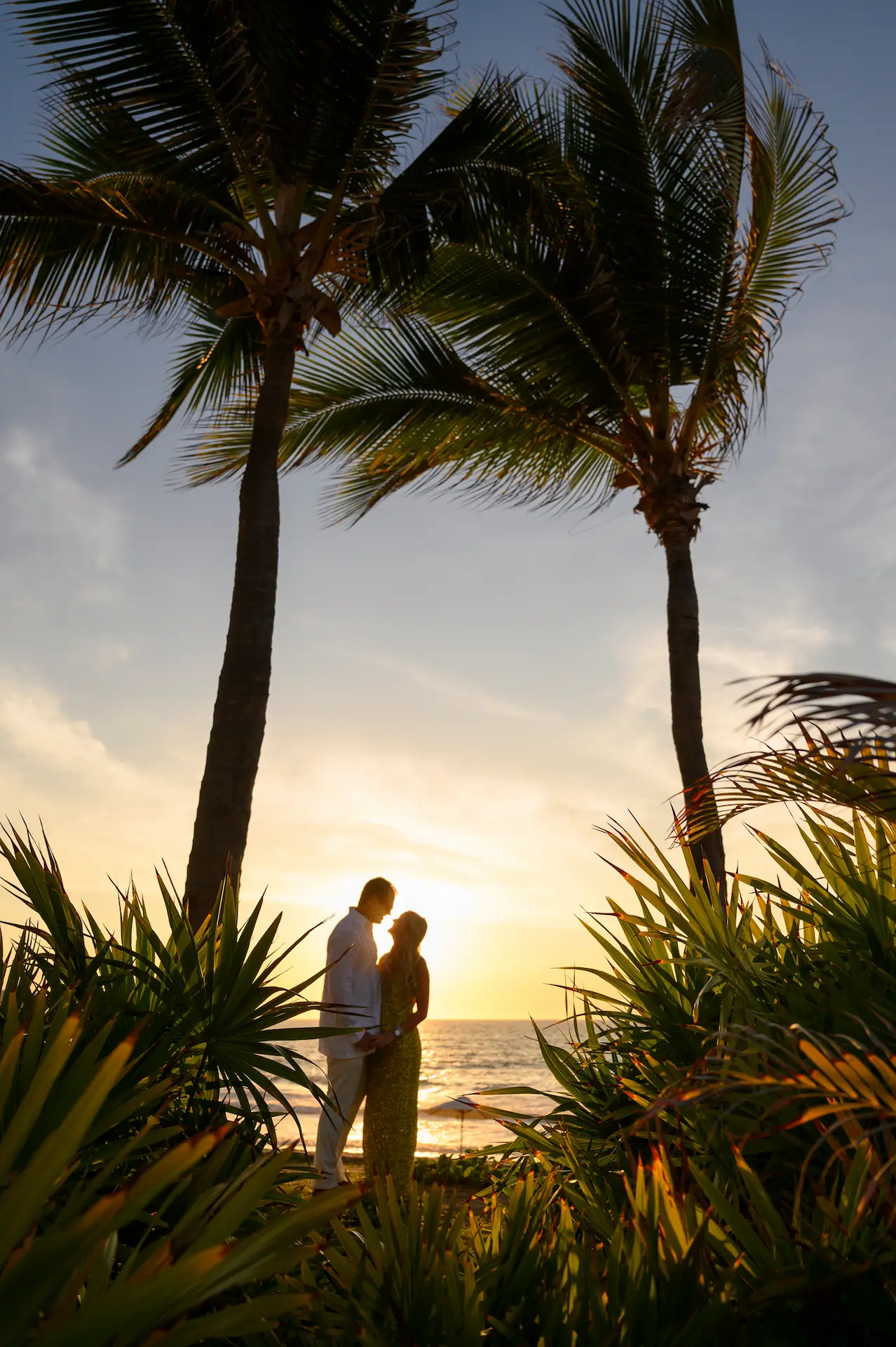 Pareja abrazándose entre palmeras al atardecer, rodeada de follaje tropical junto al océano.