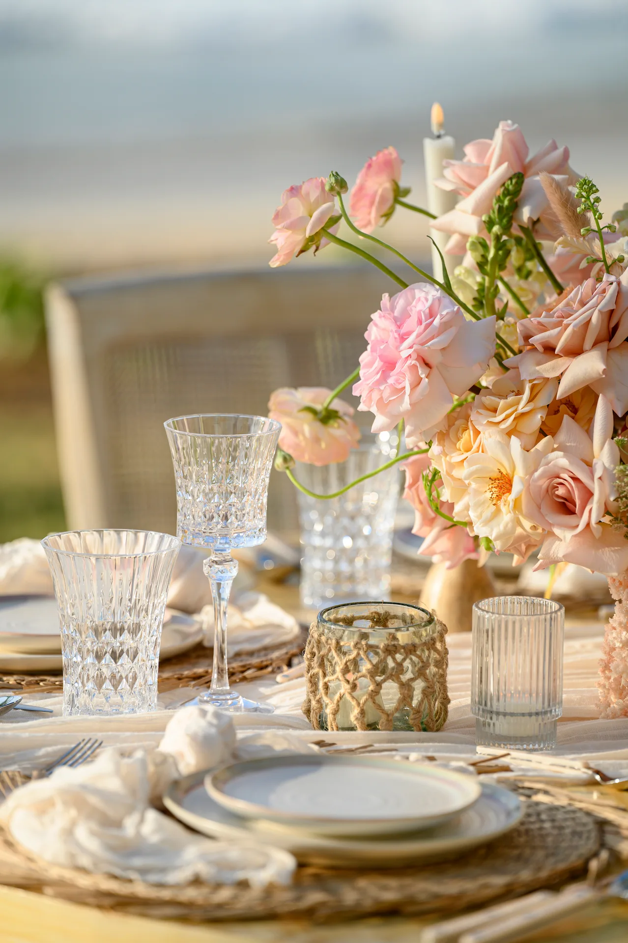 Elegante mesa junto a la playa con centro de mesa floral en tonos pastel, cristalería y manteles individuales tejidos para un evento romántico.