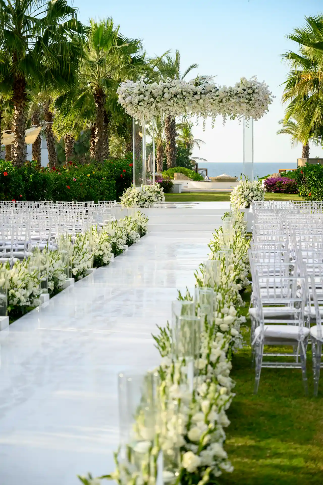Pasillo blanco brillante con flores blancas en cascada, velas de vidrio y arco floral frente al mar para una boda tropical.