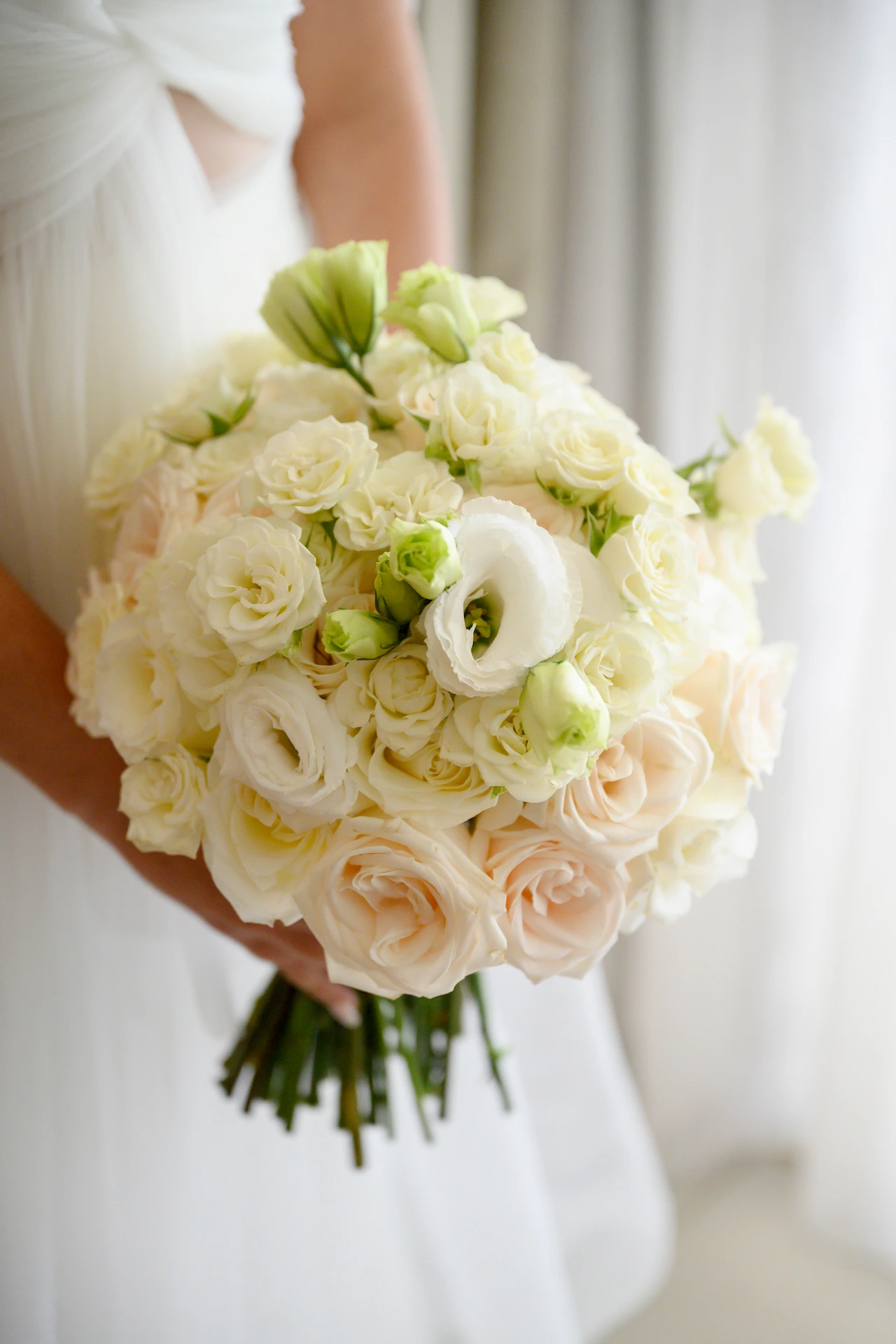 Novia sosteniendo un ramo redondo de rosas color crema y rubor, lisianthus blancos y capullos verdes.