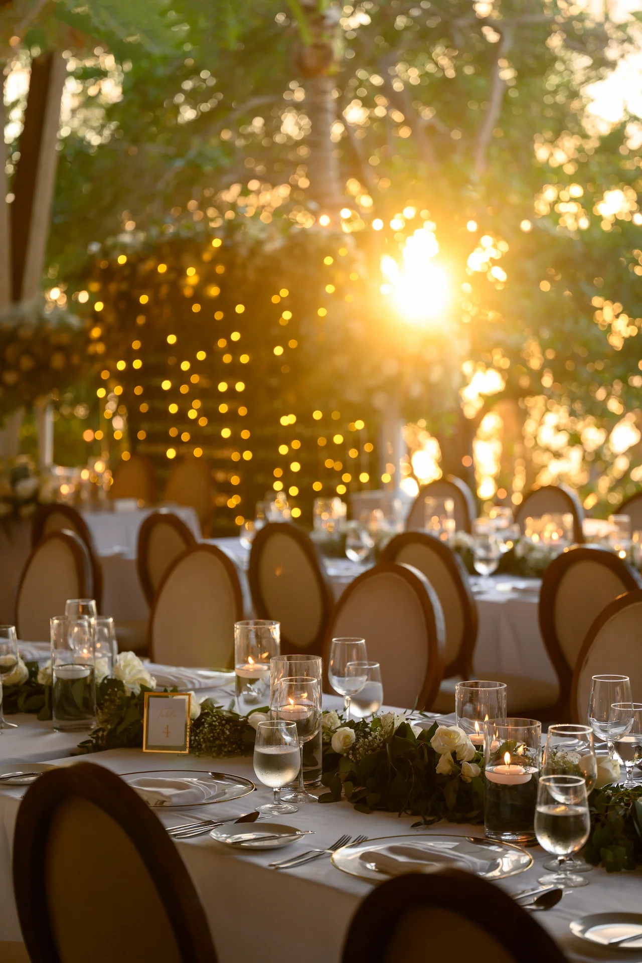 Elegante mesa de playa con mantelería blanca, rosas pastel, velas flotantes y vistas al mar al atardecer.