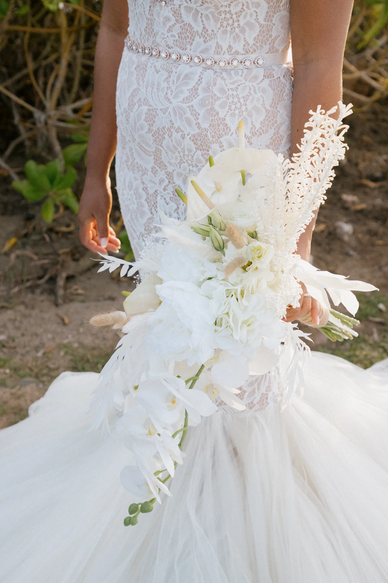Primer plano de la novia con un vestido de encaje sin tirantes sosteniendo la falda, con el anillo de bodas visible y una suave vegetación de fondo.