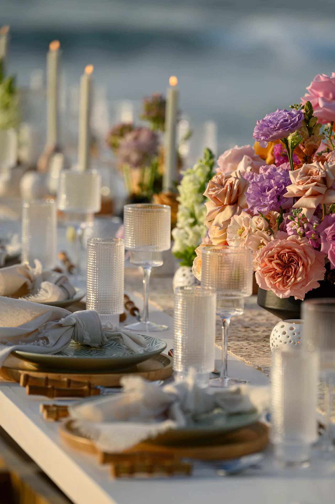 Elegante mesa junto al mar con rosas pastel, platos de cerámica, copas de cristal y velas preparadas para una celebración formal.