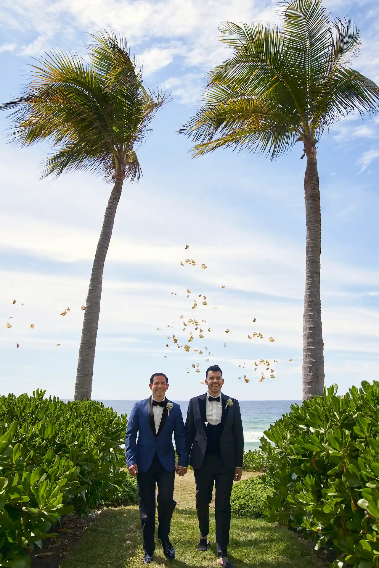 Dos novios caminando de la mano entre palmeras mientras los pétalos de las flores caen en una soleada celebración frente al mar.