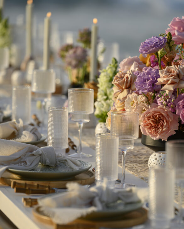 Bride and groom stand on glossy white aisle with cascading white florals and candles, framed by oceanfront floral arch at sunset.