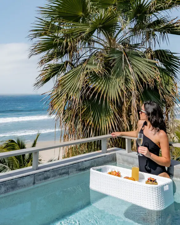 Woman relaxing in rooftop plunge pool with floating breakfast, champagne, and panoramic ocean views.