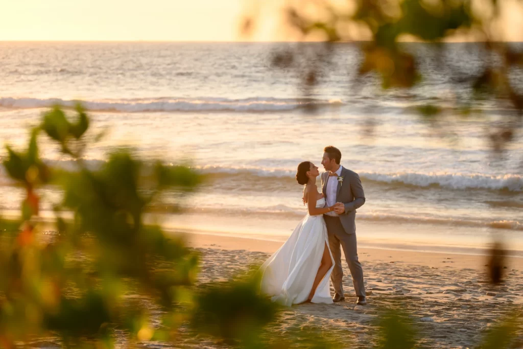 Bride and groom embracing on beach at sunset with ocean waves and golden light in the background.